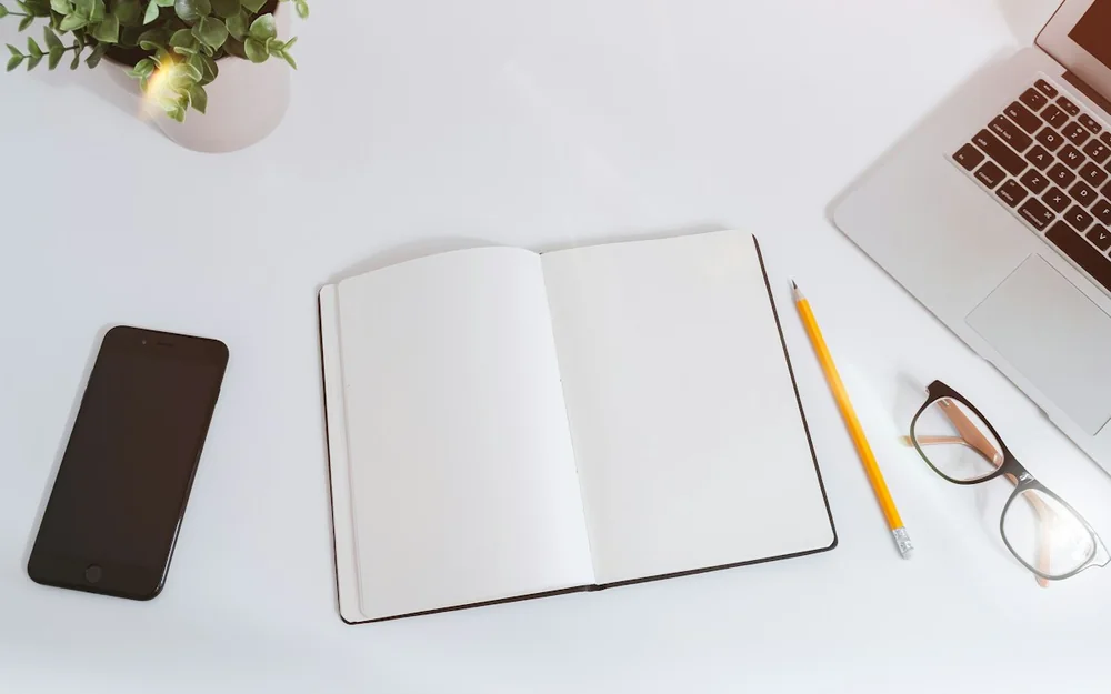 An open notebook, pencil and laptop arranged on a white desk.